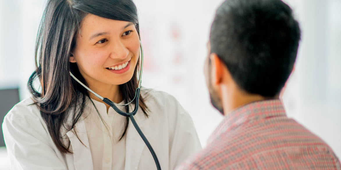 Female healthcare provider smiling while examining a male patient with a stethoscope.
