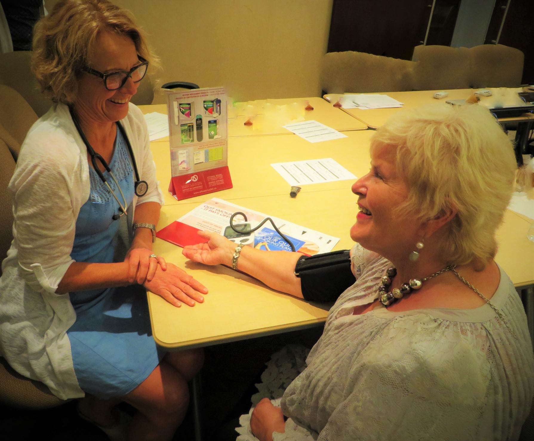 A healthcare provider sitting across an elderly woman getting her blood pressure taken, engaging in pleasant conversation.
